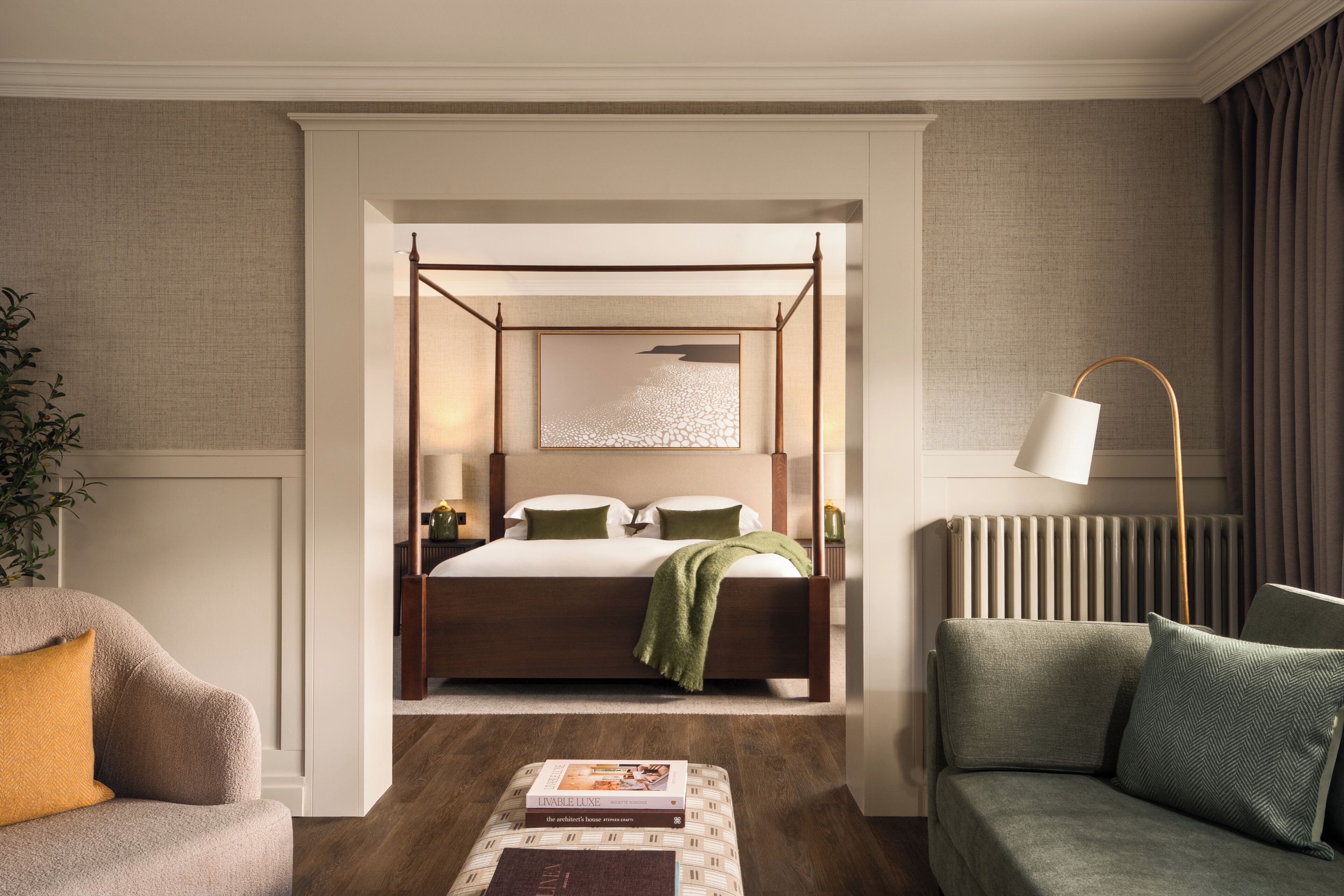 A hotel bedroom with a four-poster bed, a bedside table with a lamp, a green throw blanket, a sofa, and a coffee table with books.