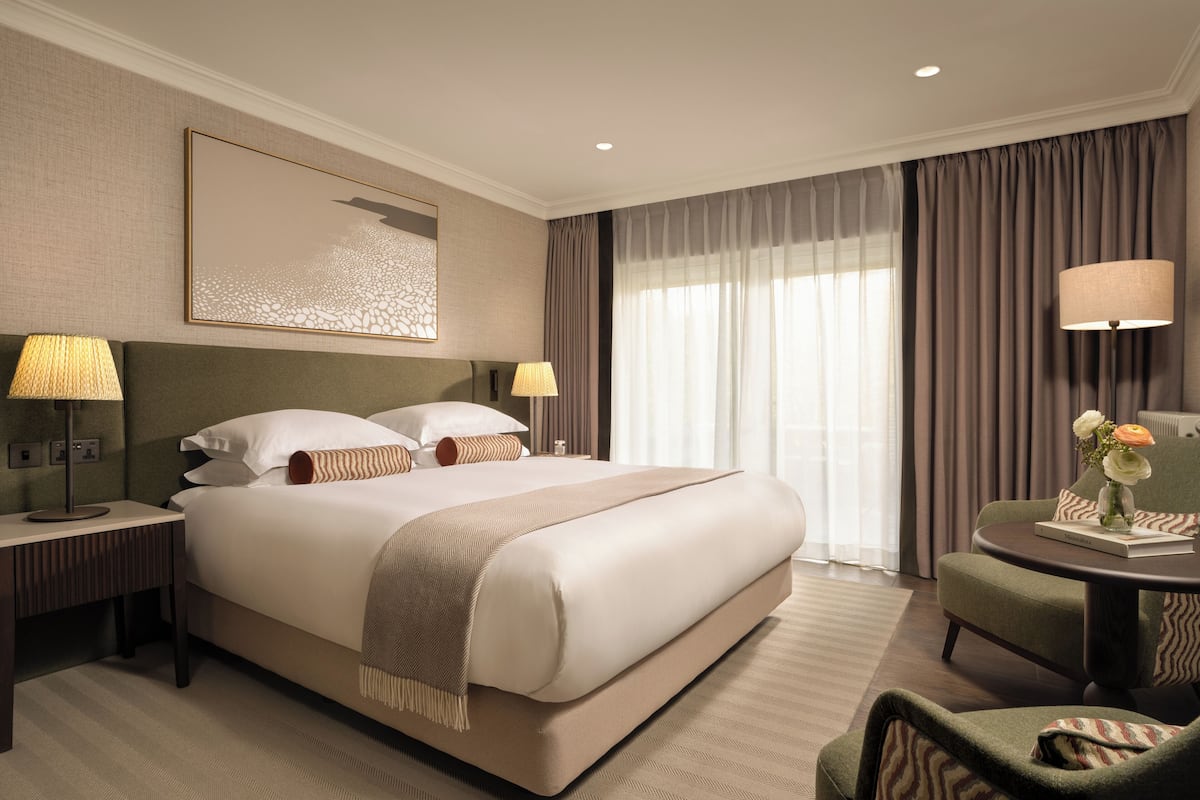 A hotel room with a large bed, bedside lamps, a chair, a table with a vase of flowers, and a painting on the wall.
