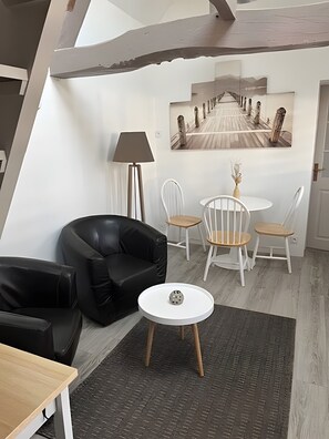 A modern room with a black leather sofa, a round white table with a decorative item, a lamp, and a painting of a bridge. - Gîte “Le Bois Flotté” in Nature with Wi-Fi (Boismont)