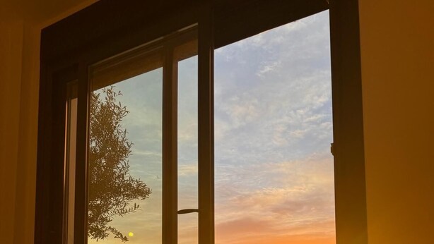 Ein Zimmer mit großem Fenster, einem Bett und Blick auf den Sonnenuntergang.