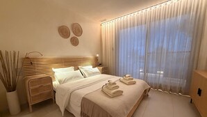 Ein ordentlich bezogenes Bett mit weißen Leinen, ein Nachttisch mit Lampe und ein großes Fenster mit durchsichtigen Vorhängen.