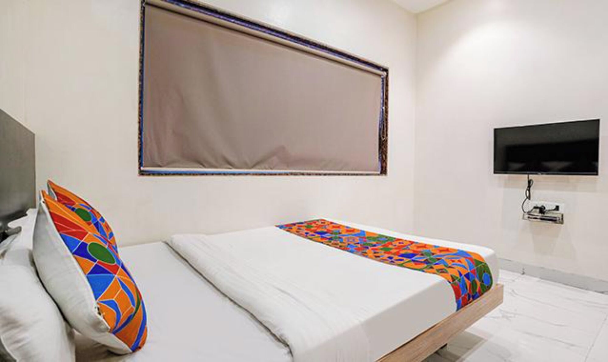 Un lit simple avec un oreiller coloré et une couverture unie, un téléviseur fixé au mur et une fenêtre avec un store.
