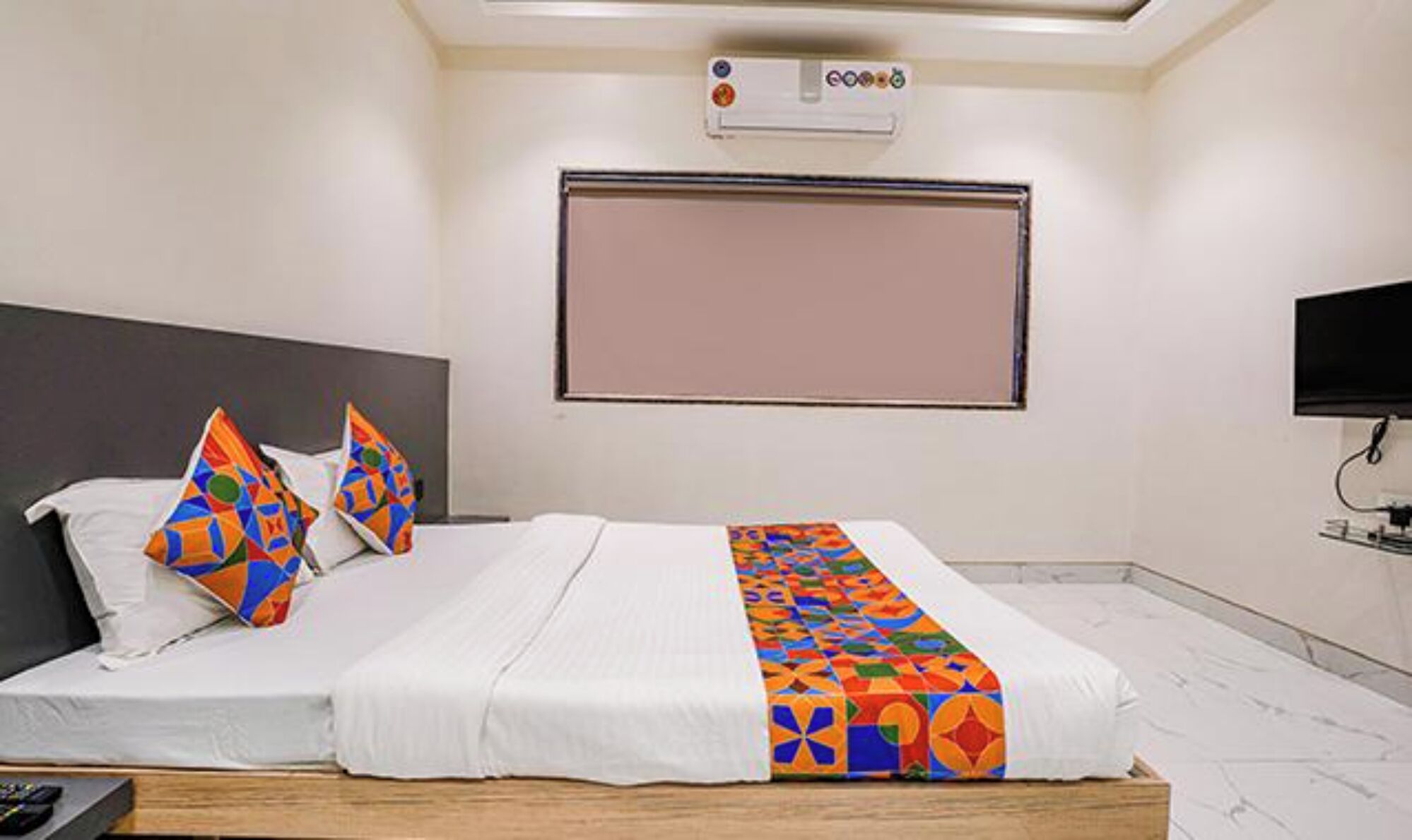 Una habitación de hotel con cama, almohadas, manta, televisor y aire acondicionado.