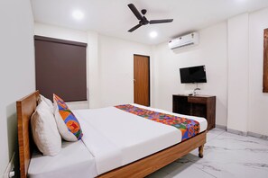 Habitación de hotel con cama, ventilador de techo, unidad de aire acondicionado, televisor y mesita de noche de madera.