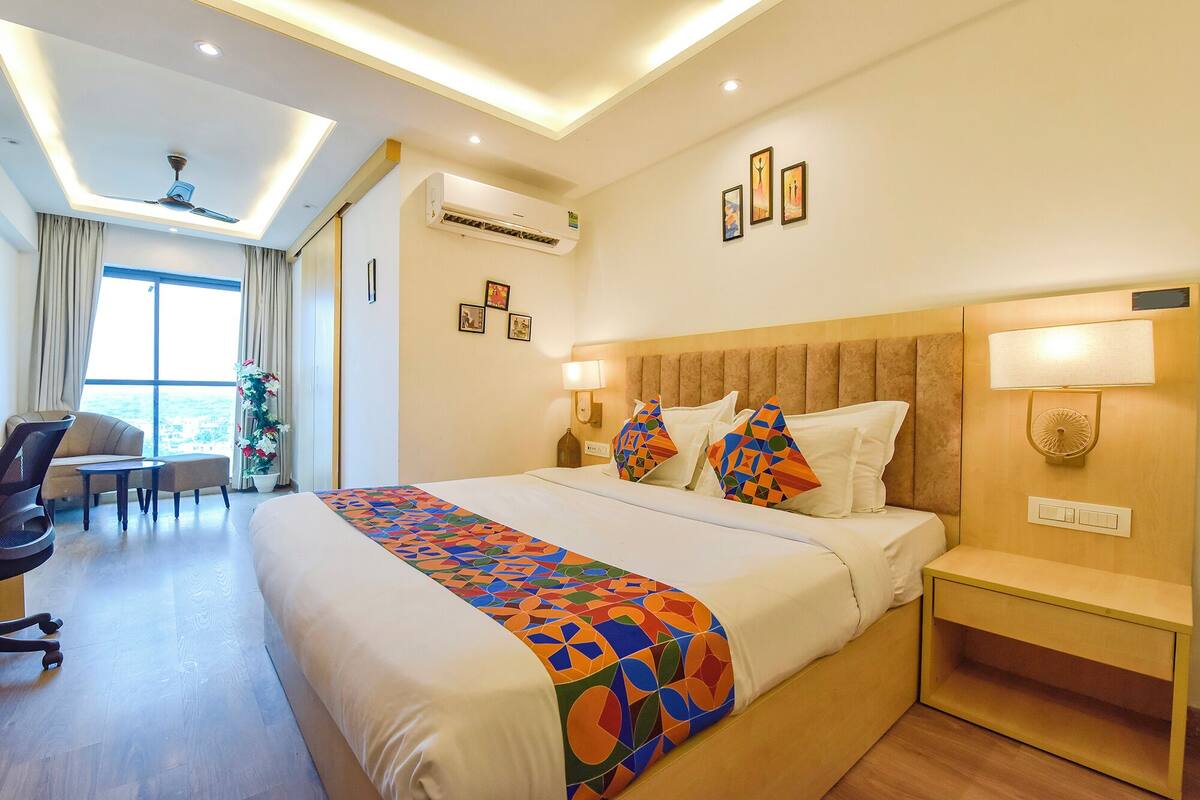 A hotel room with a bed, bedside tables, a desk, a chair, an air conditioning unit, and a window with curtains.