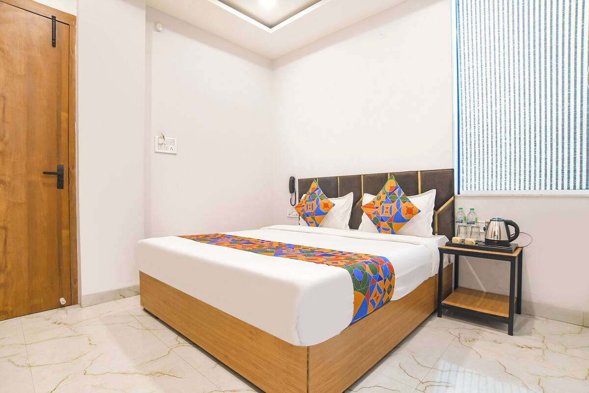 Habitación de hotel con cama de madera, ropa de cama blanca, un plaid geométrico de colores, una mesita de noche con hervidor y una ventana con persianas.