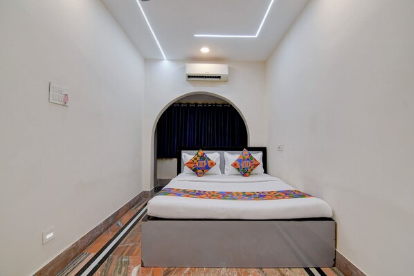 A narrow hotel room with a bed, an air conditioning unit, and a small window with a curtain.