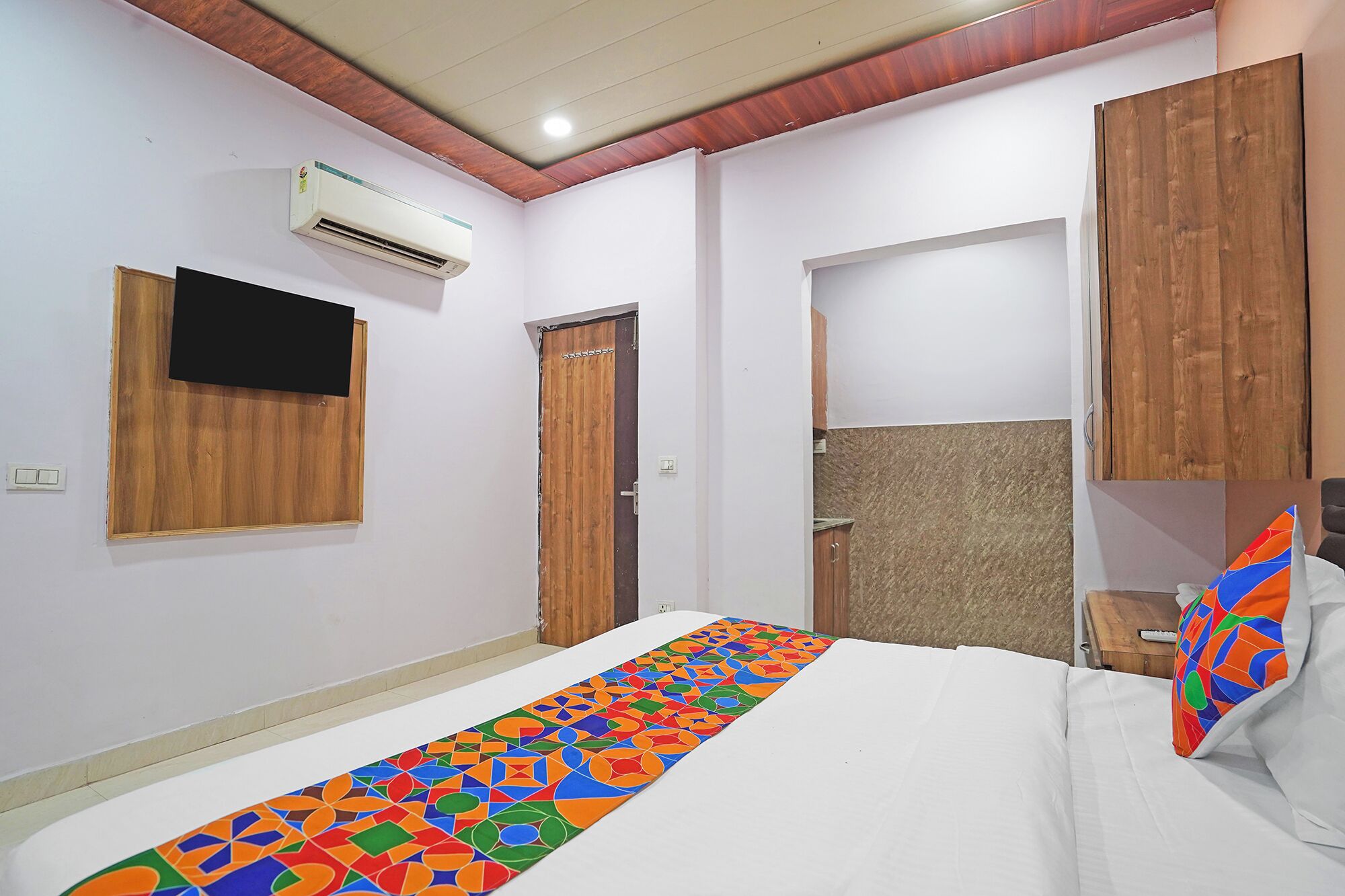 A bedroom with a bed, a TV mounted on the wall, an air conditioning unit, wooden wardrobe, and a colorful rug.