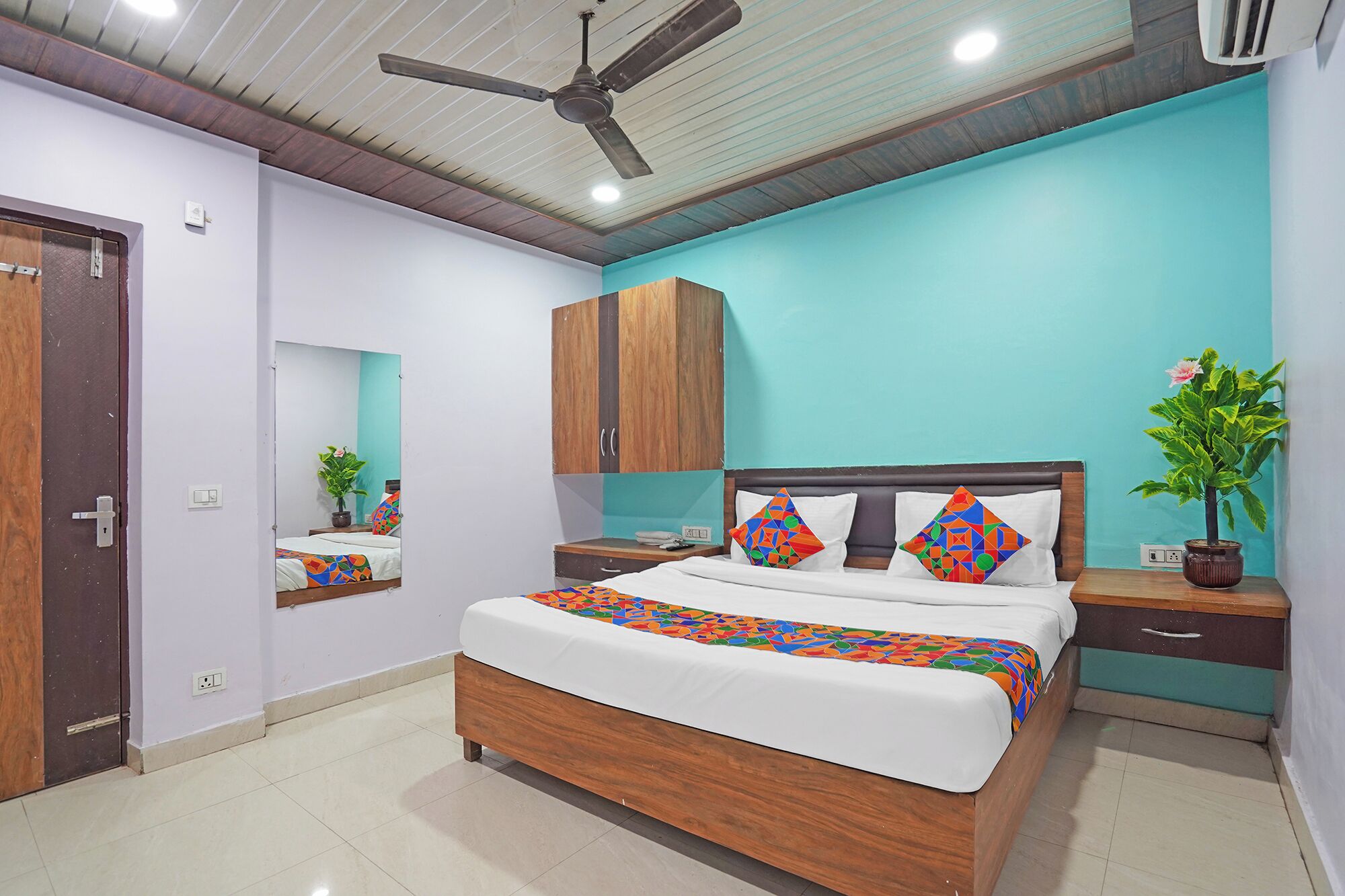 A bedroom with a bed, a ceiling fan, a wooden wardrobe, a nightstand, a potted plant, and a mirror.