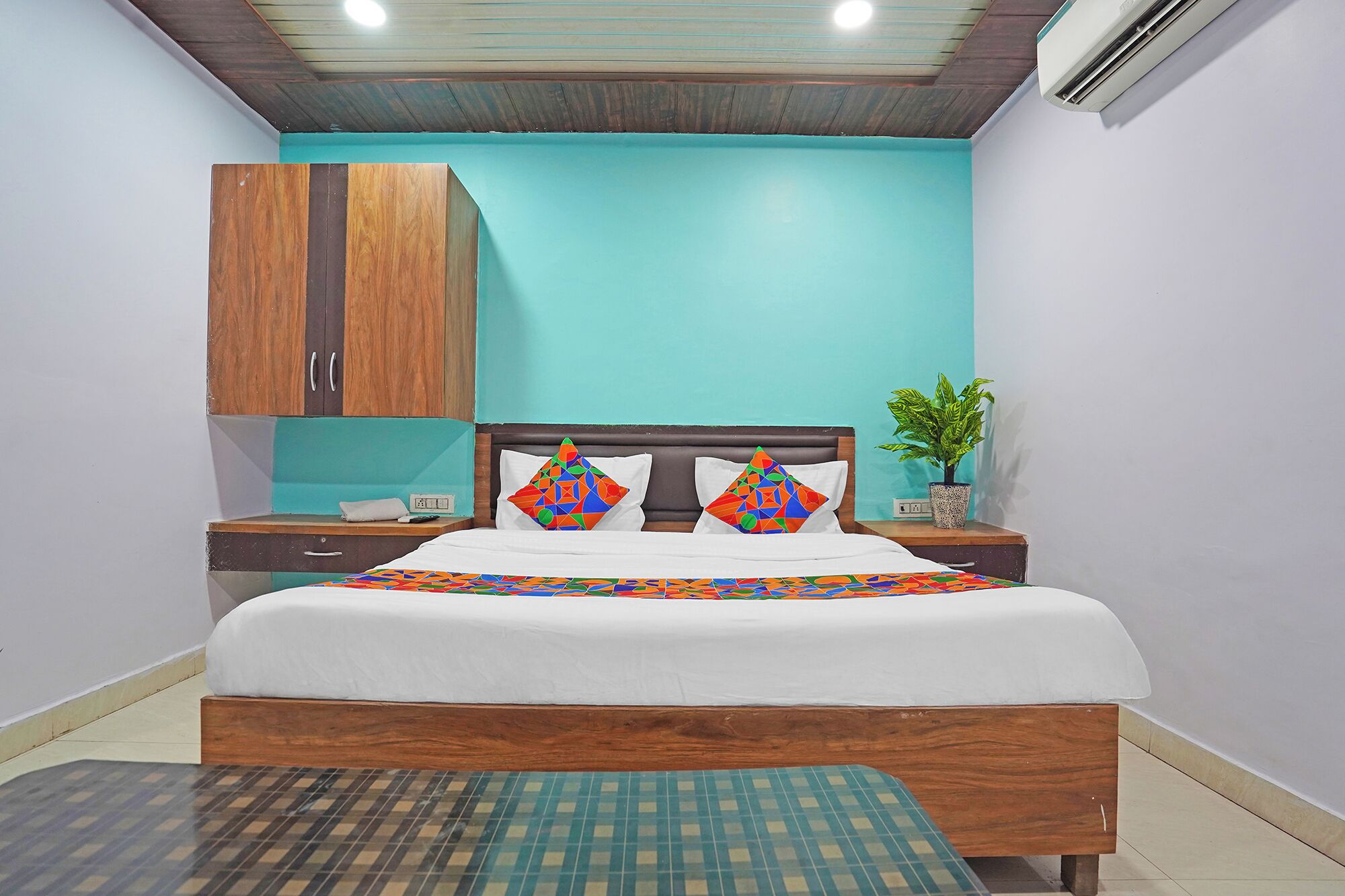 A bedroom with a bed, a nightstand, a wooden wardrobe, a potted plant, and a tiled floor.