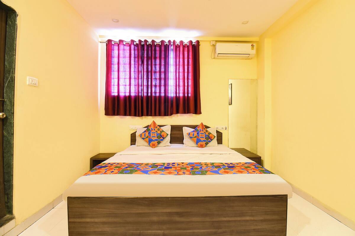 A narrow room with a bed, a window with curtains, an air conditioning unit, and a door.