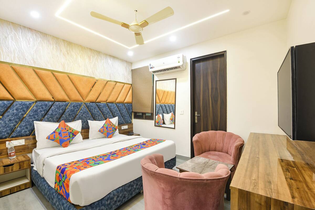 A hotel room with a bed, a pink armchair, a wooden table, a ceiling fan, and an air conditioning unit.