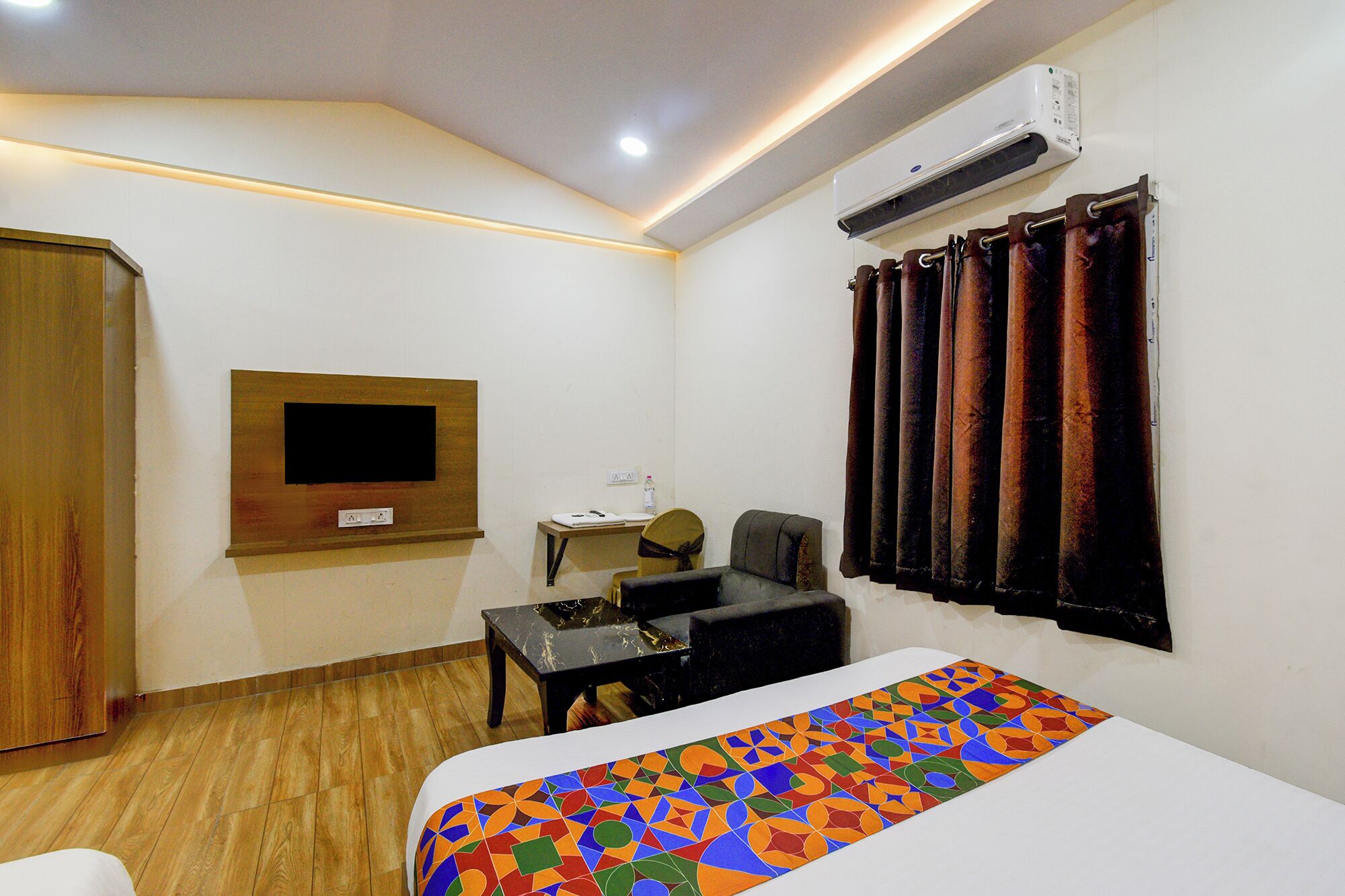 A hotel room with a bed, a desk, a chair, a TV mounted on the wall, and an air conditioning unit.