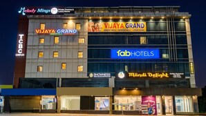 Un hotel de varios pisos con letreros que indican 'Vijaya Grand', 'Fab Hotels' y 'Mandi Delight'.