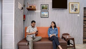 A brown sofa with a person on the left and a child on the right, a small stool, and a wall with framed pictures and a clock. - Fabhotel Comfort Zone (Mumbai)