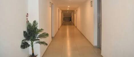 A long, narrow hotel corridor with a potted plant on the left, doors on both sides, and a green exit sign on the right.
