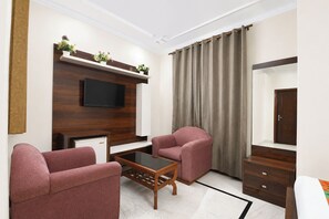 A living room with a flat-screen TV, two armchairs, a coffee table, a cabinet, and a large window with curtains. - Fabhotel The Arjun (Gurugram)