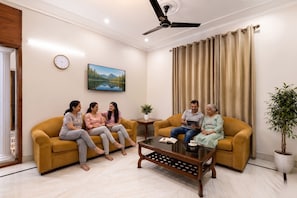 A living room with a sofa, a coffee table, a clock, a painting, and a ceiling fan. - FabHotel The Arjun Sushant lok - Iffco Chowk Sector 29 (Gurugram)