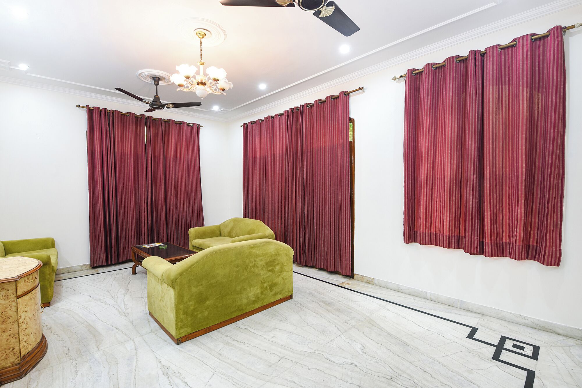 Una habitación con ventilador de techo, candelabro, cortinas rojas, sofá verde y suelo de mármol.