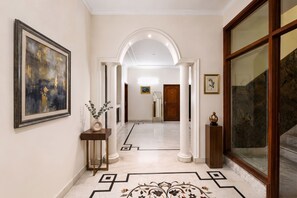 A hallway with a marble floor, framed artwork, a vase with plants, and a staircase with a glass railing. - Fabhotel The Arjun (Gurugram)