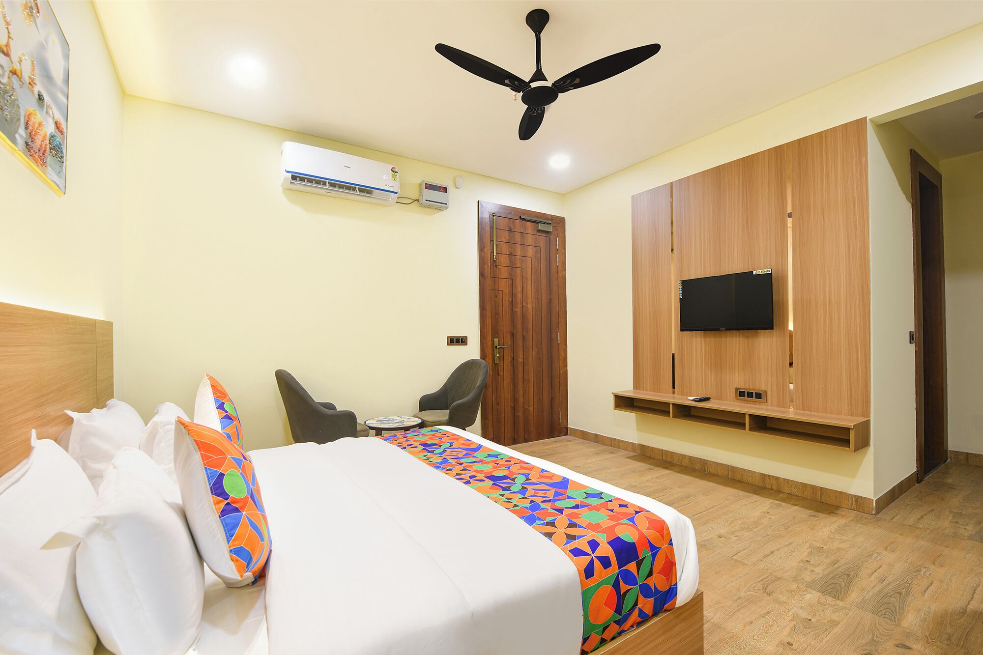 A hotel room with a bed, a ceiling fan, an air conditioning unit, a TV mounted on the wall, and a wooden wardrobe.