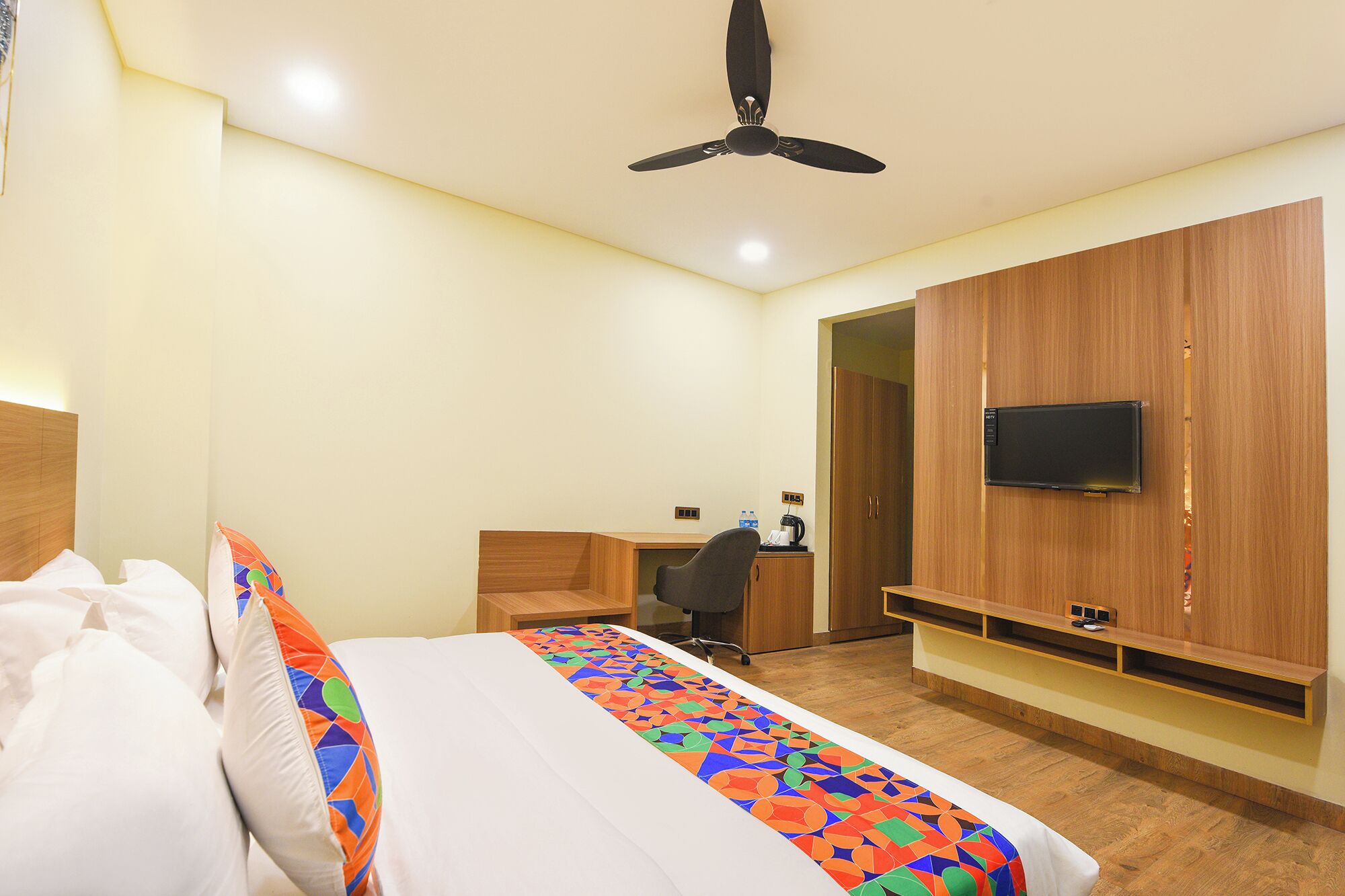 Habitación de hotel con una cama, un escritorio, una silla, un televisor montado en la pared y un ventilador de techo.
