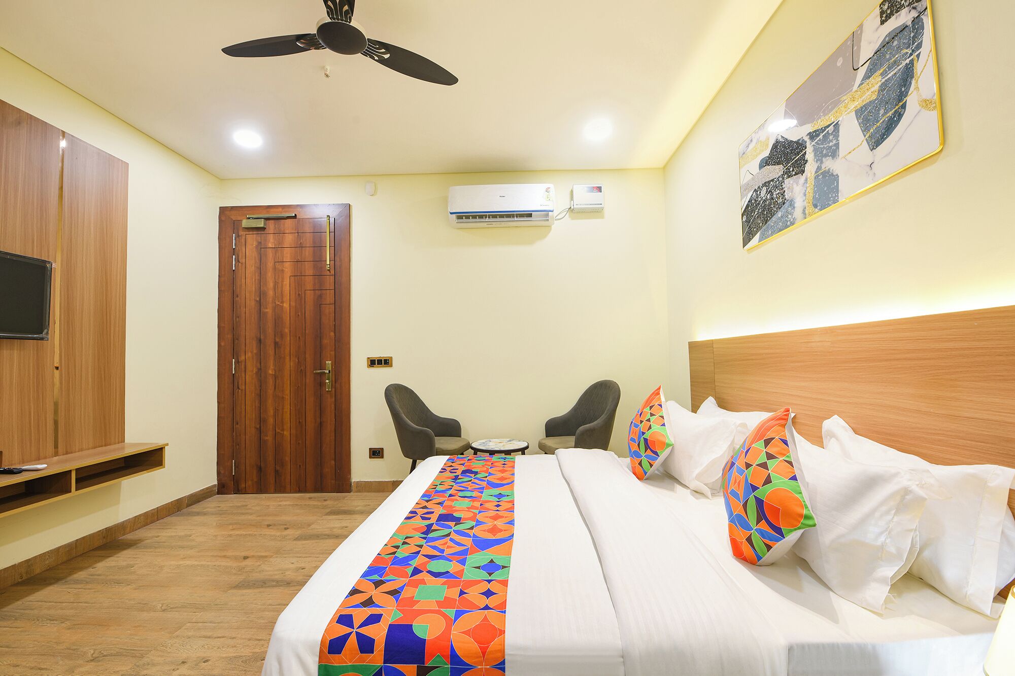 A hotel room with a bed, two chairs, a wooden wardrobe, an air conditioning unit, and a ceiling fan.