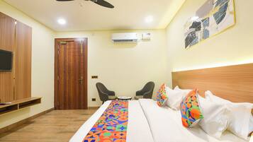 A hotel room with a bed, two chairs, a wooden wardrobe, an air conditioning unit, and a ceiling fan.