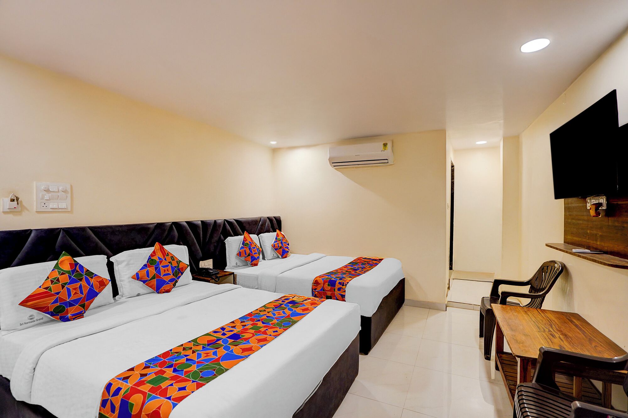 A hotel room with two beds, a wooden table, a chair, an air conditioning unit, and a television mounted on the wall.