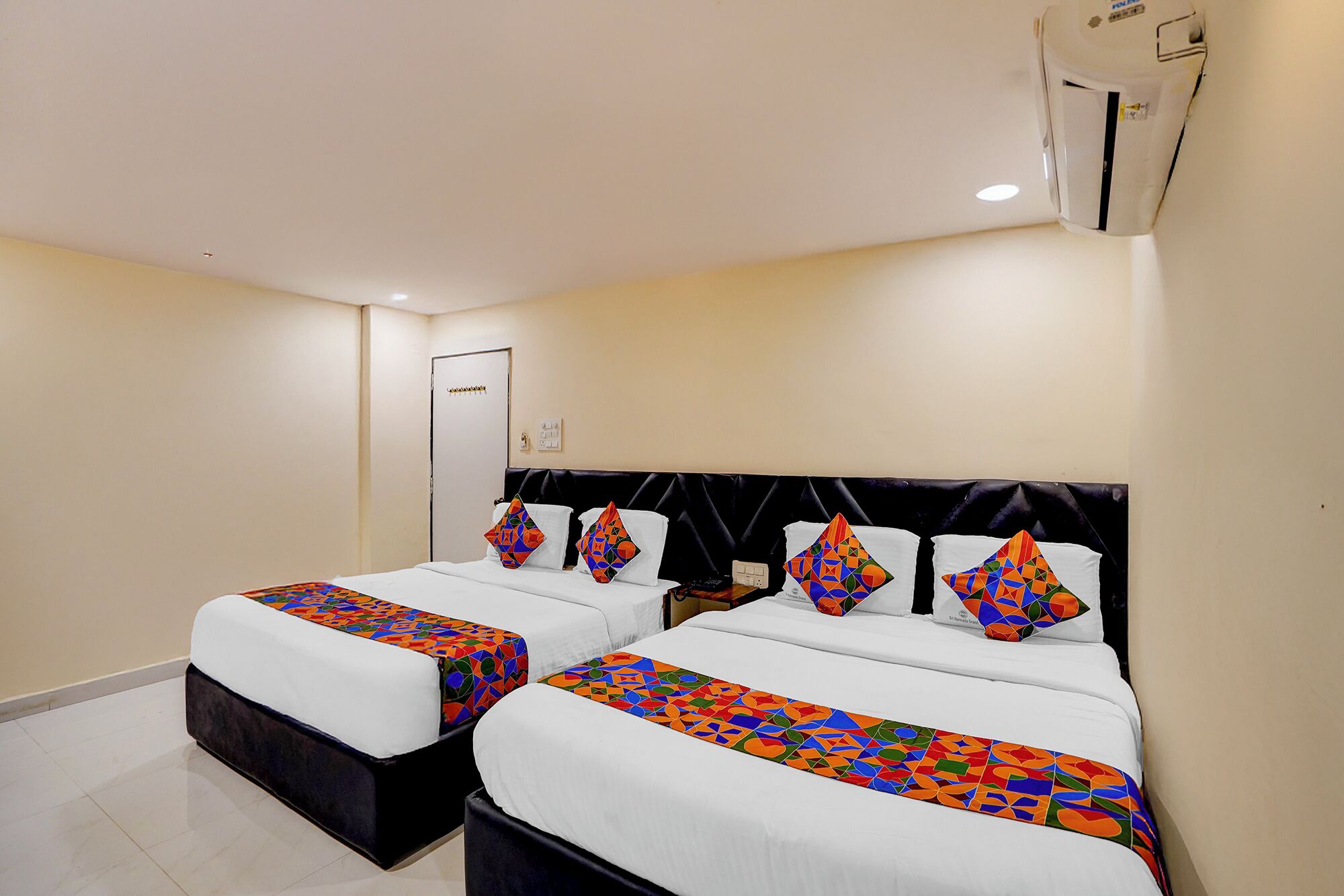 A hotel room with two beds, each with a black headboard and white bedding. There is a door with a handle and a light switch on the wall.