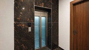 Um elevador com porta de vidro, uma porta de madeira à direita e uma parede de mármore com padrão salpicado.