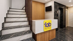 Bancada de recepção de hotel com a placa 'Tab Hotels', escadaria com degraus de mármore, painel de parede de madeira e parede de mármore preto, e um elevador.