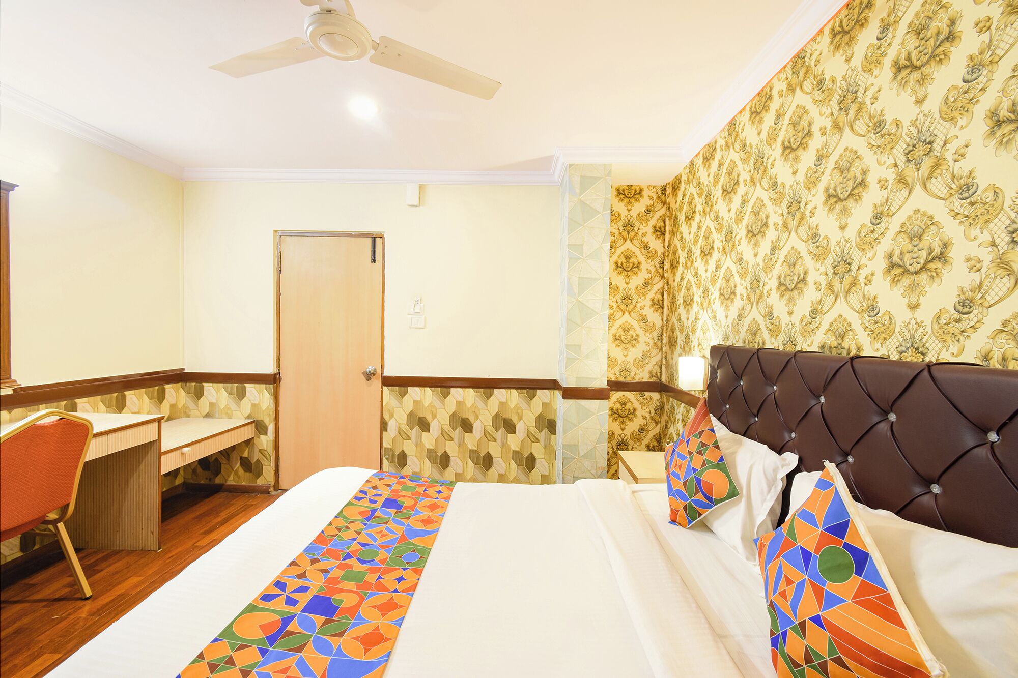 A hotel room with a bed, a desk, a chair, a ceiling fan, and patterned wallpaper.