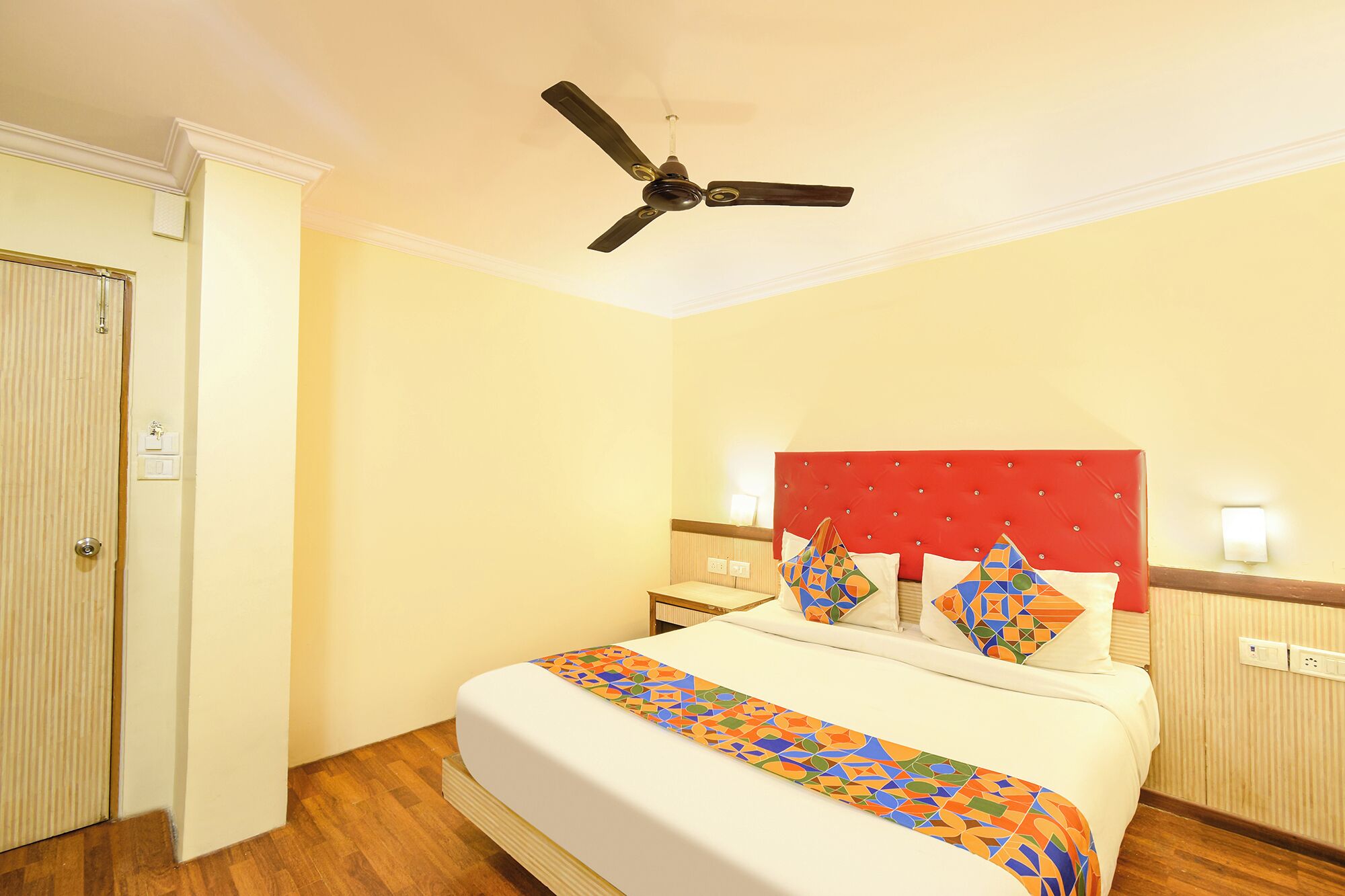 Une chambre d’hôtel avec un lit, un ventilateur de plafond, une tête de lit rouge et un plancher en bois.