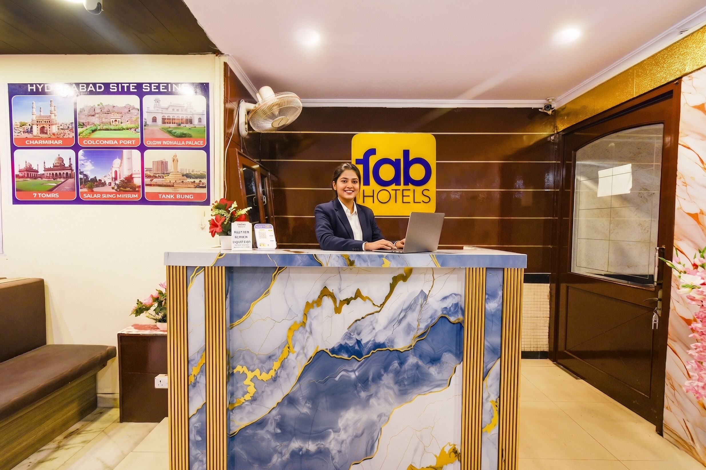 A hotel reception desk with a laptop, a sign for 'fab hotels,' and a poster of Hyderabad sites.