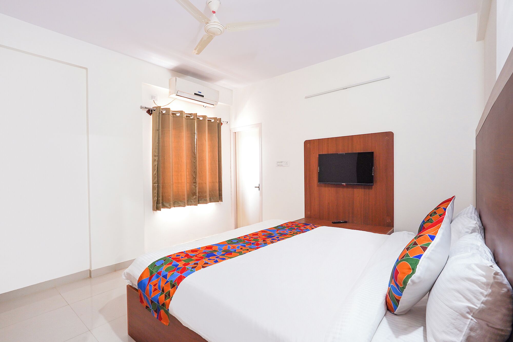 A hotel room with a bed, a TV mounted on the wall, a ceiling fan, and a window with curtains.