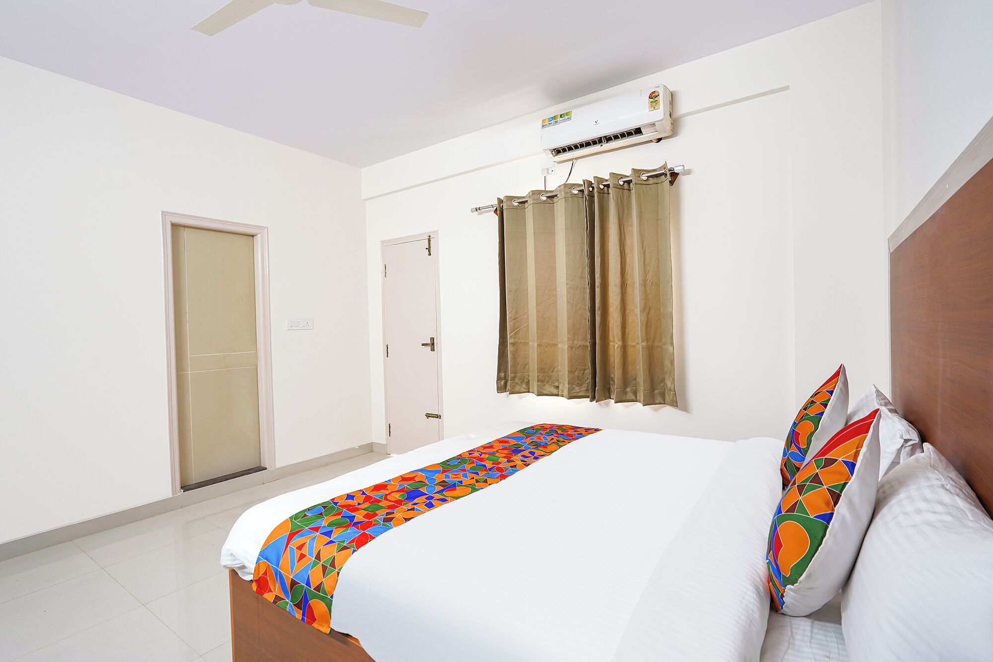 A hotel room with a bed, a ceiling fan, an air conditioning unit, a window with curtains, and a door.