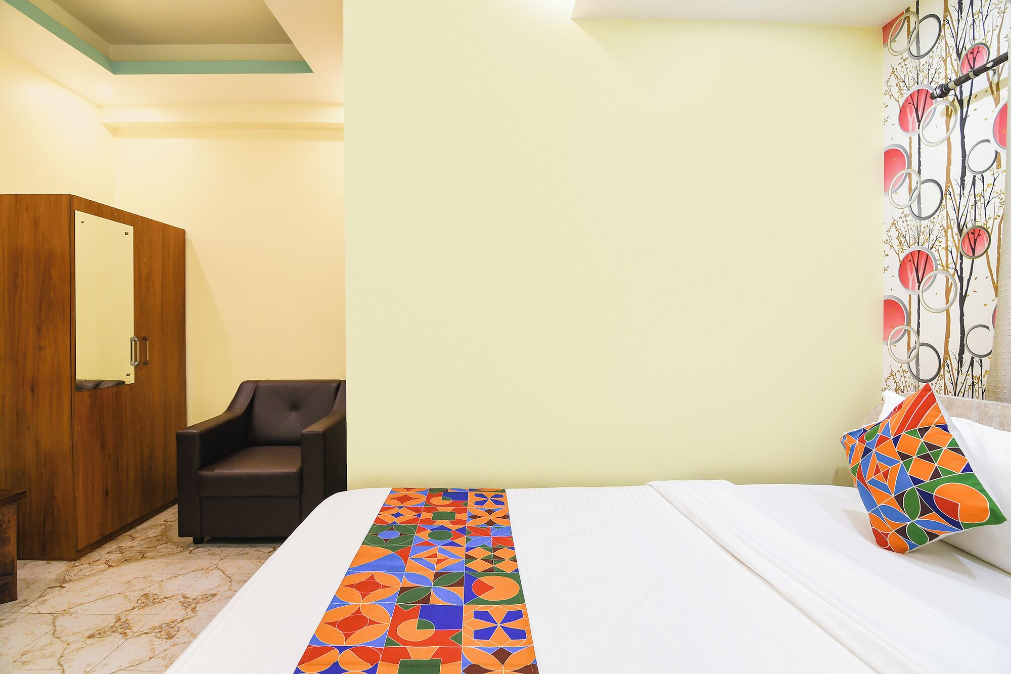 Une chambre d’hôtel avec un lit, une chaise, une armoire en bois et un mur orné d’un miroir et d’un rideau à motifs.