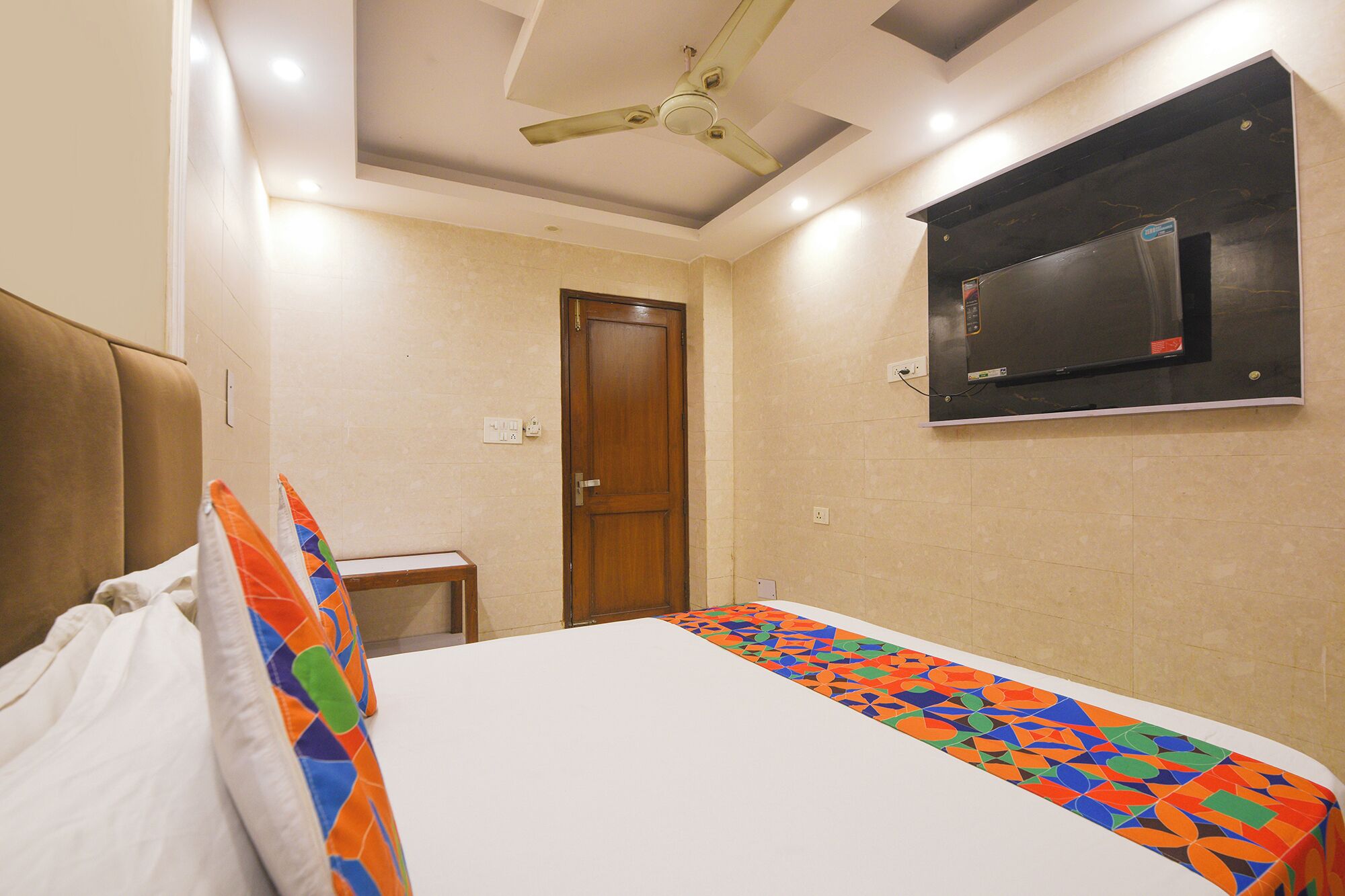 A hotel room with a bed, a ceiling fan, a TV mounted on the wall, and a door.