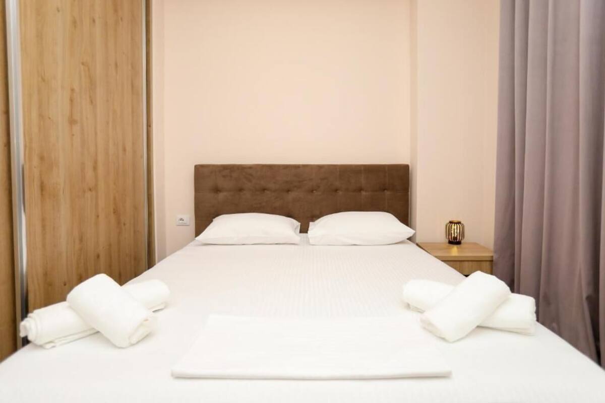 Una cama bien hecha con sábanas y almohadas blancas, un cabecero de madera y una mesita de noche con una lámpara.