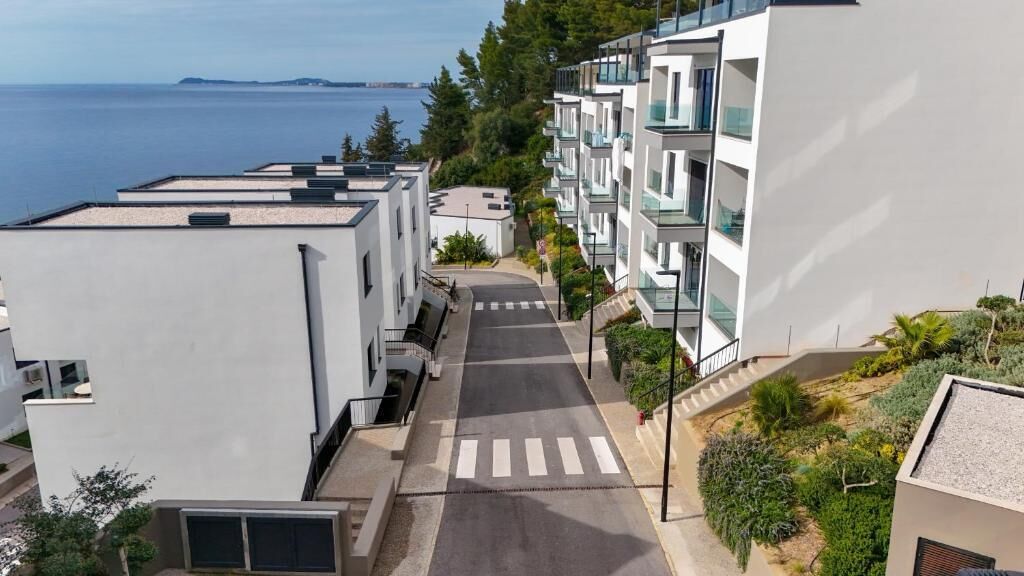 Un quartier résidentiel moderne, avec une route menant à un immeuble doté de balcons, entouré de verdure et offrant une vue sur la mer.