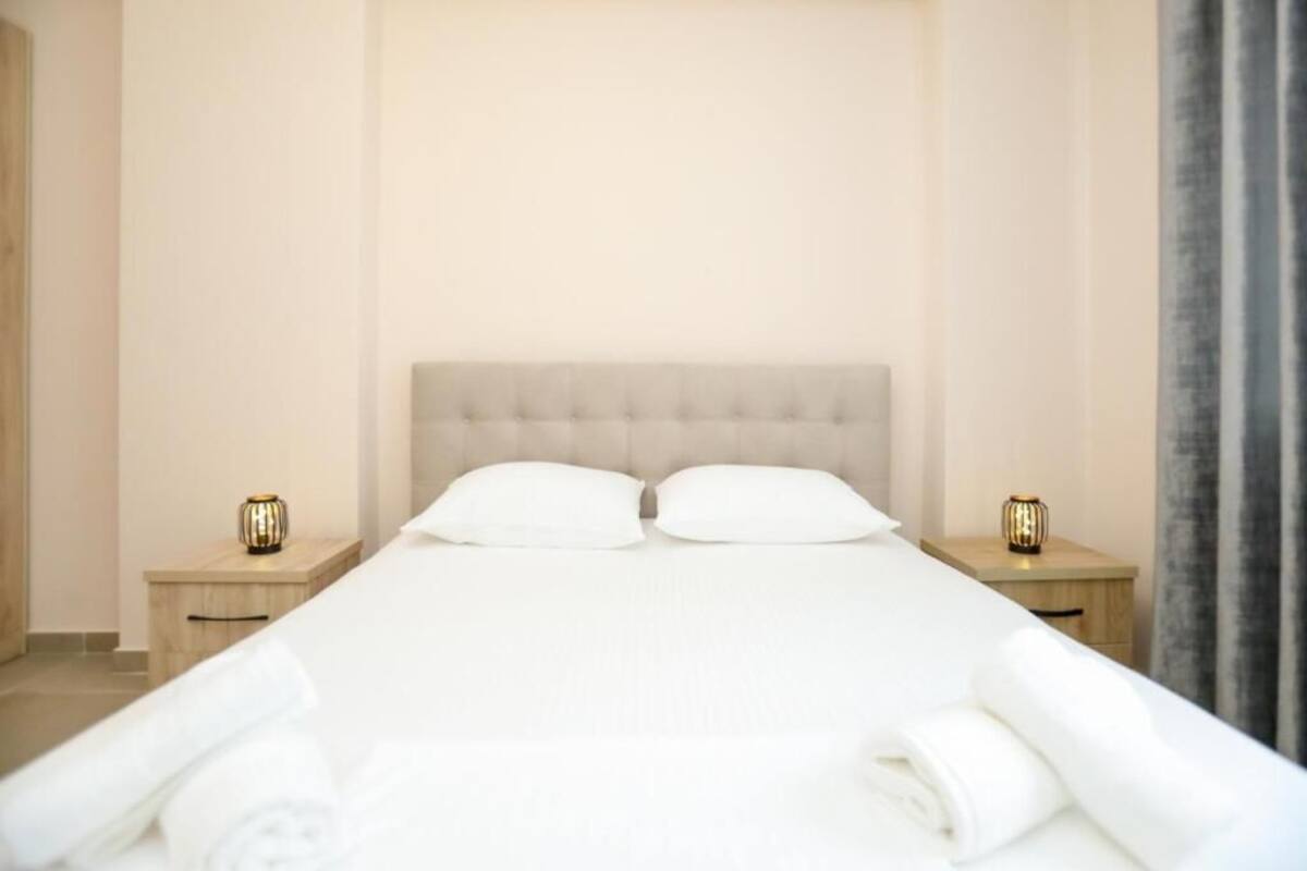Una cama bien hecha con cabecera capitonada, dos mesitas de noche con lámparas decorativas y una pared de color neutro.
