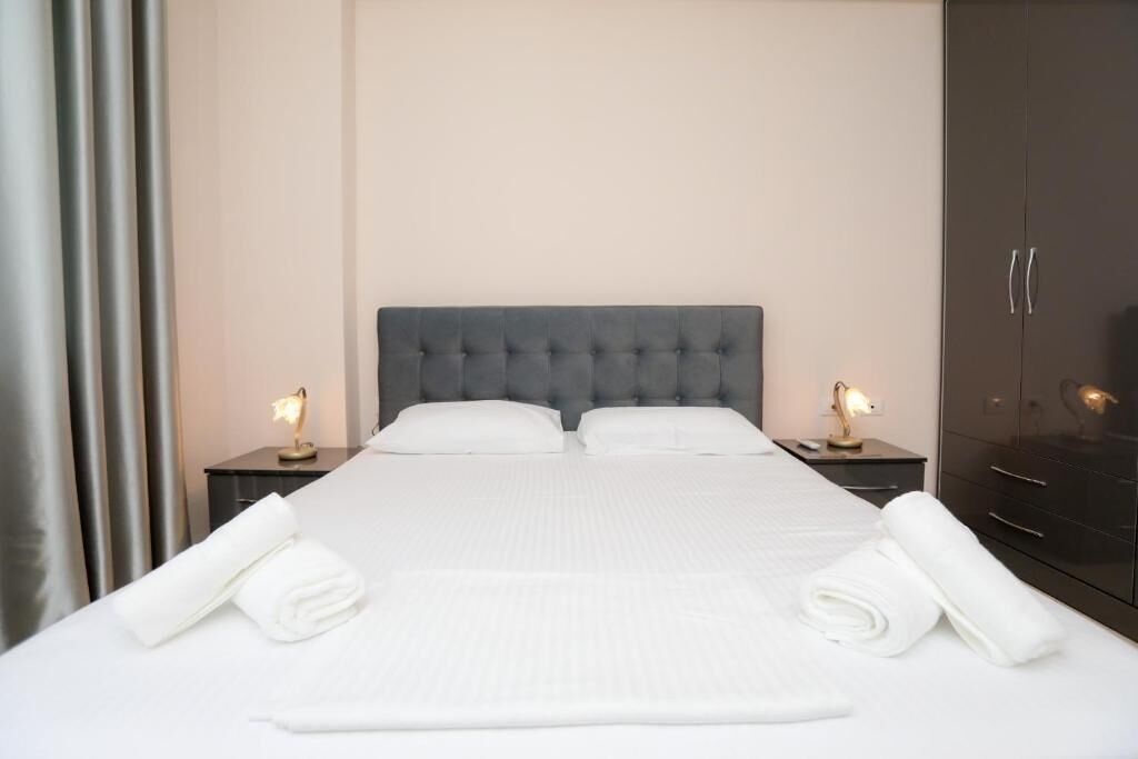 Een net opgemaakt bed met witte lakens en twee opgerolde handdoeken, een grijze gestuufde hoofdbord, nachtlampjes en een donkere garderobe.