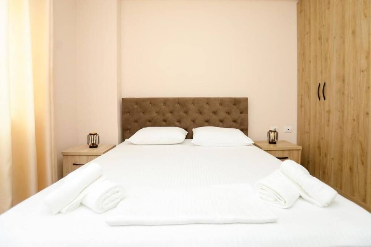Una cama bien tendida con un cabecero capitonado, dos mesitas de noche con jarrones decorativos y un armario de madera.