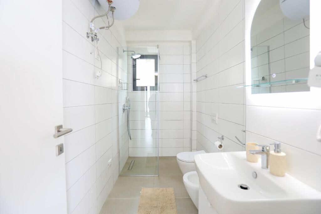 Un baño moderno con azulejos blancos, mampara de vidrio, inodoro e lavamanos con espejo.