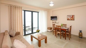 A living room with a sofa, a coffee table, a dining table with chairs, a TV, and a sliding glass door leading to an outdoor area. - Velaris Apartments (Vlore)