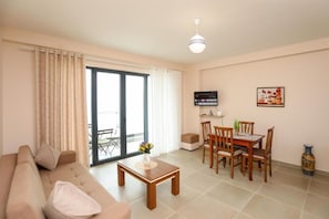 A living room with a sofa, a coffee table, a dining table with chairs, a TV, and a sliding glass door leading to an outdoor area. - Velaris Apartments (Vlore)