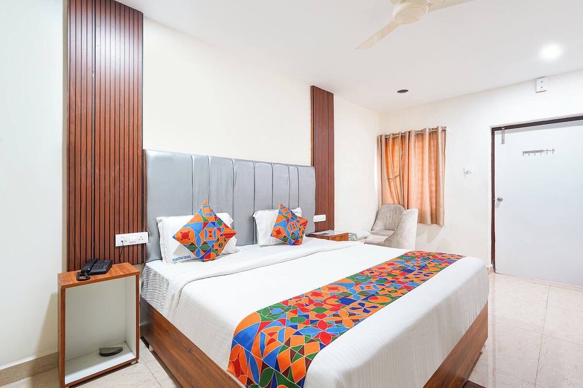 A hotel room with a bed, a bedside table with a phone, a chair, a door, and a window with curtains.