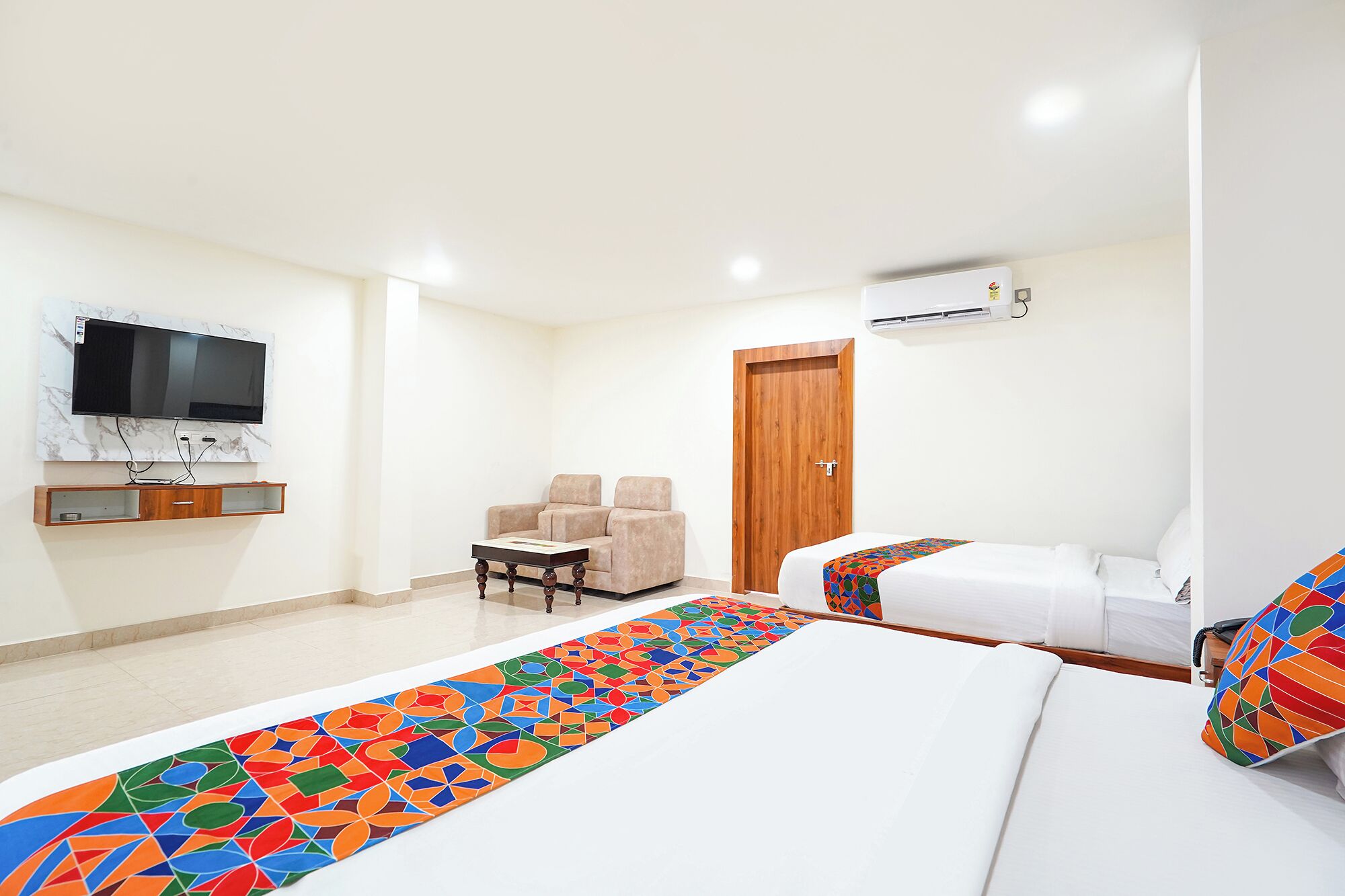 A hotel room with a TV mounted on the wall, a wooden cabinet below, a sofa, a bed with a colorful blanket, and an air conditioning unit.