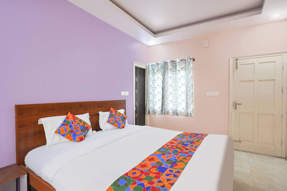 A bedroom with a wooden headboard, a bed with white bedding, colorful pillows, a patterned bedspread, a window with floral curtains, and a closed door.